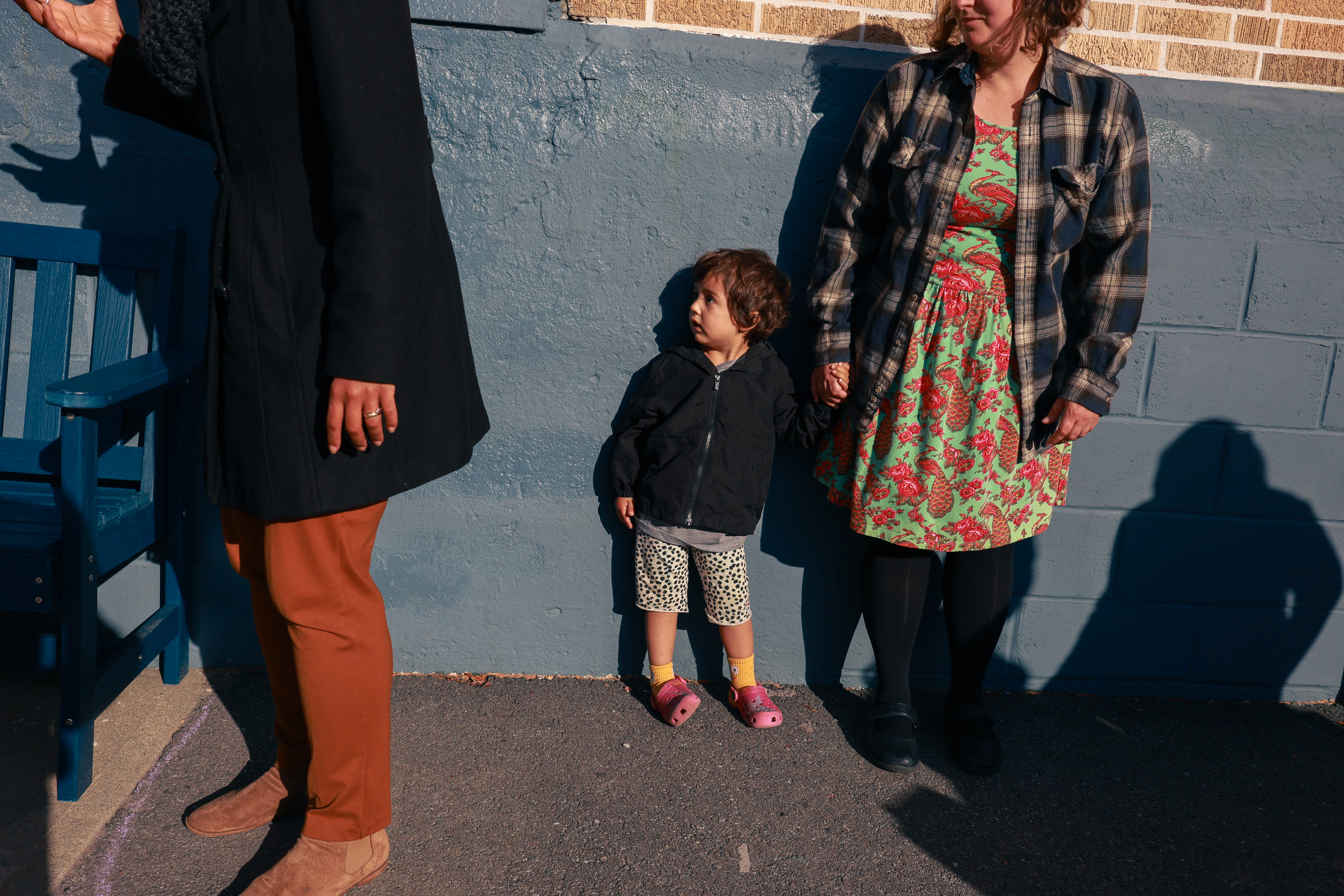 A small child wearing a black zip-up sweatshirt and pink crocs holds the hand of a woman in a green and pink dress. The two stand against a blue wall.
